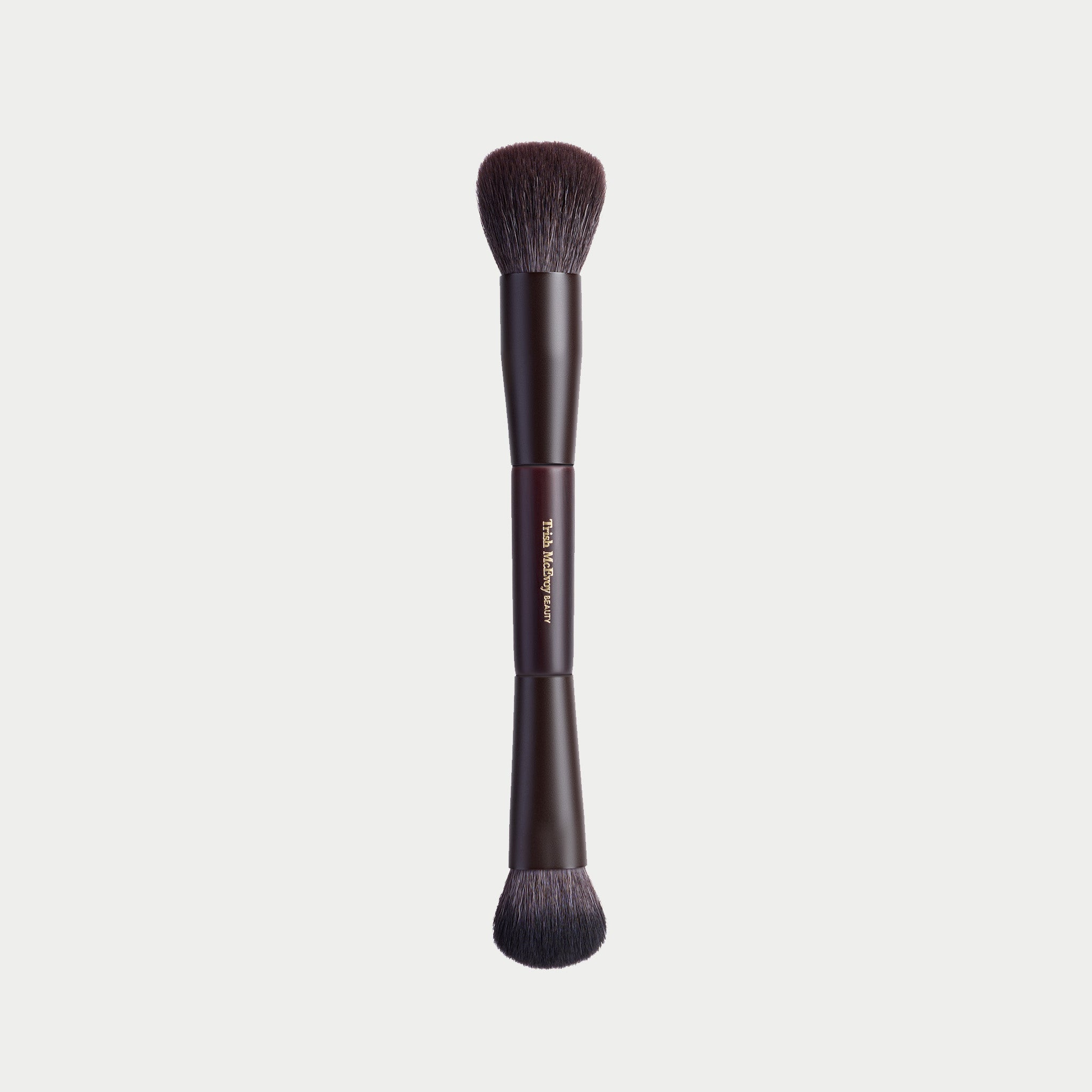 A dual-ended makeup brush with dark brown bristles and a dark brown handle, featuring the 'Tula Bejovi' brand name printed in gold on the center. The brush is centered against a solid light gray background.