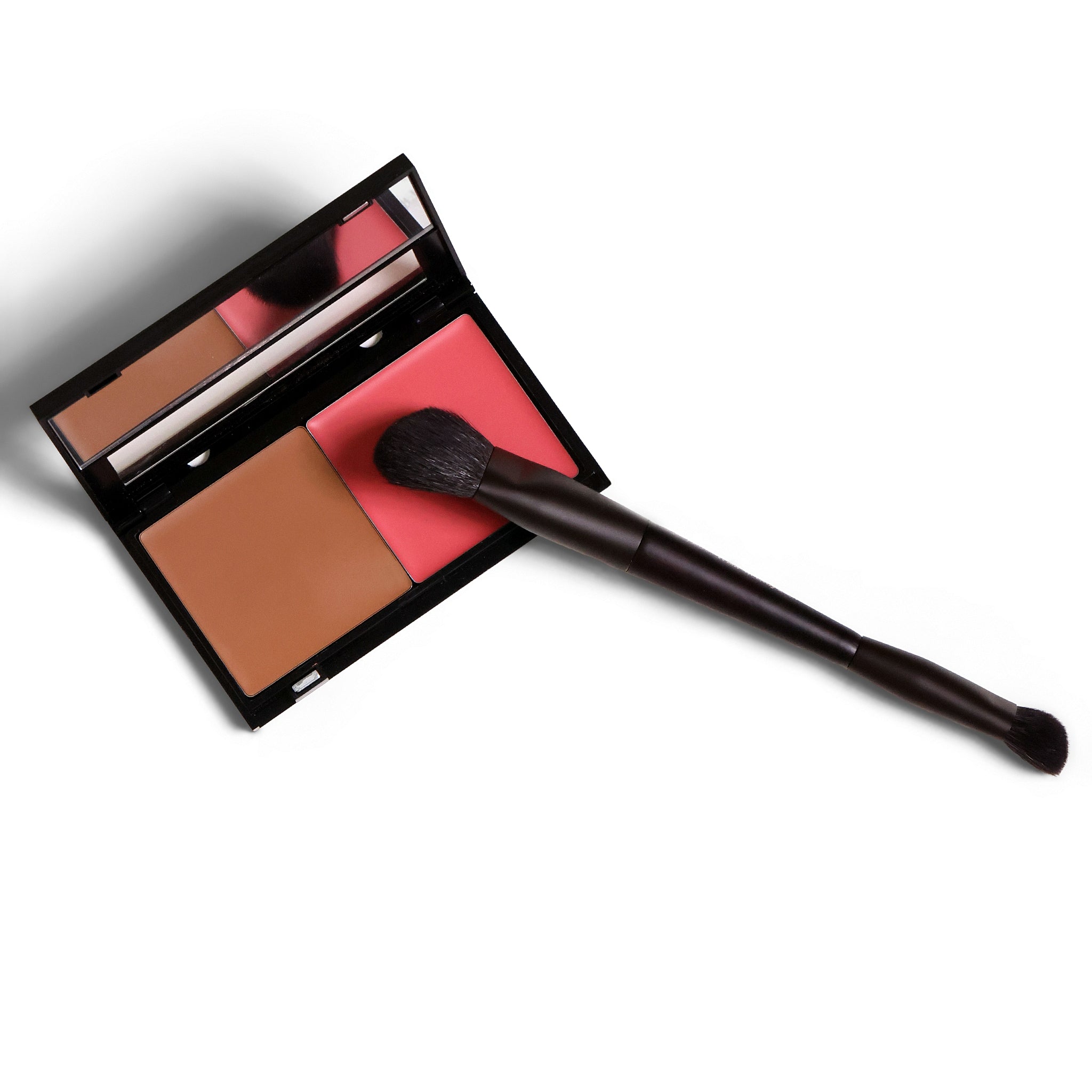 An open makeup compact with a mirror, two pressed powder pans in tan and coral pink, and a dark brown dual-ended brush resting across the compact, shown on a white background.