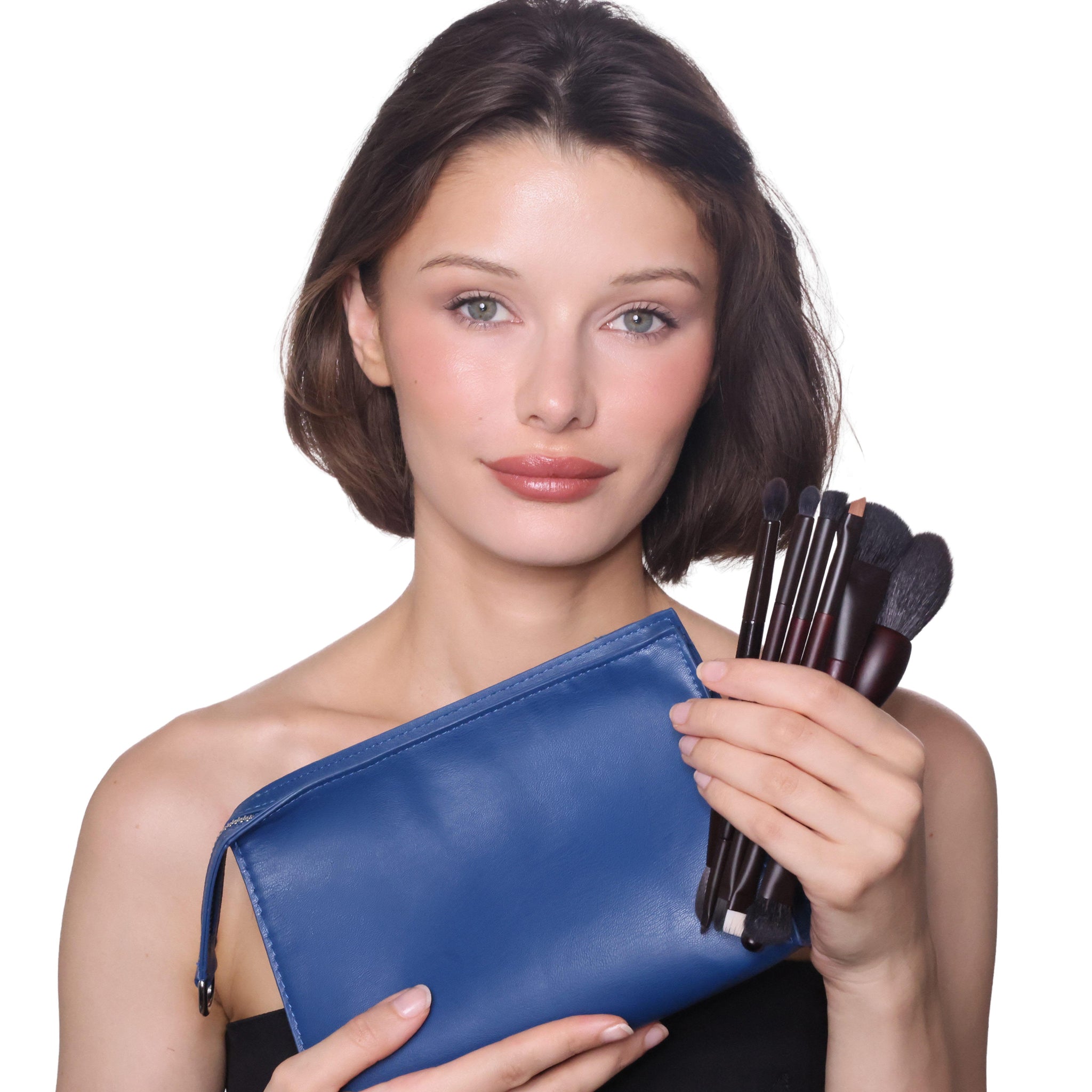 A woman with short brown hair holds a blue makeup pouch and a set of makeup brushes against a plain white background.