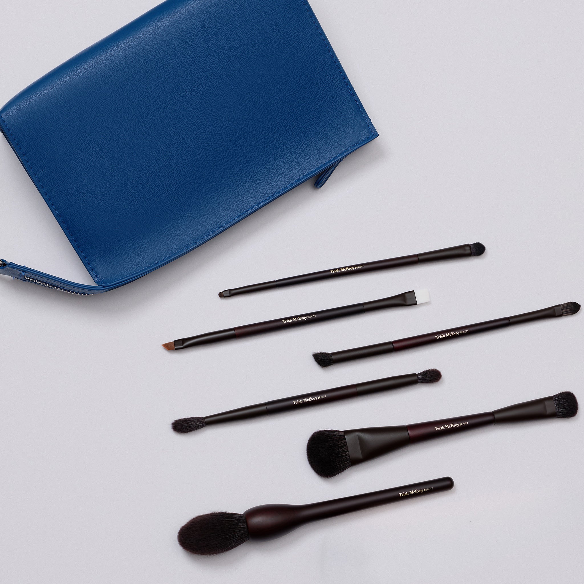 A six-piece makeup brush set with dark brown bristles and black handles, arranged next to a blue zippered pouch on a light gray background.