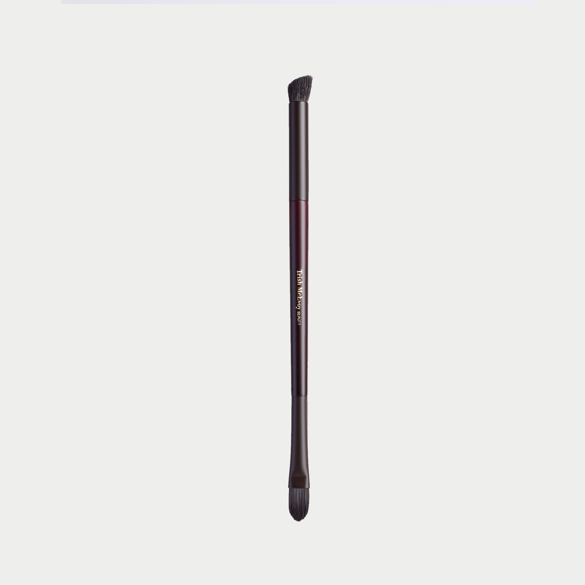 A single makeup brush with a dark metallic handle and black bristles, shown vertically against a light gray background. The handle features a metallic rose gold accent band with gold lettering that reads 'm.