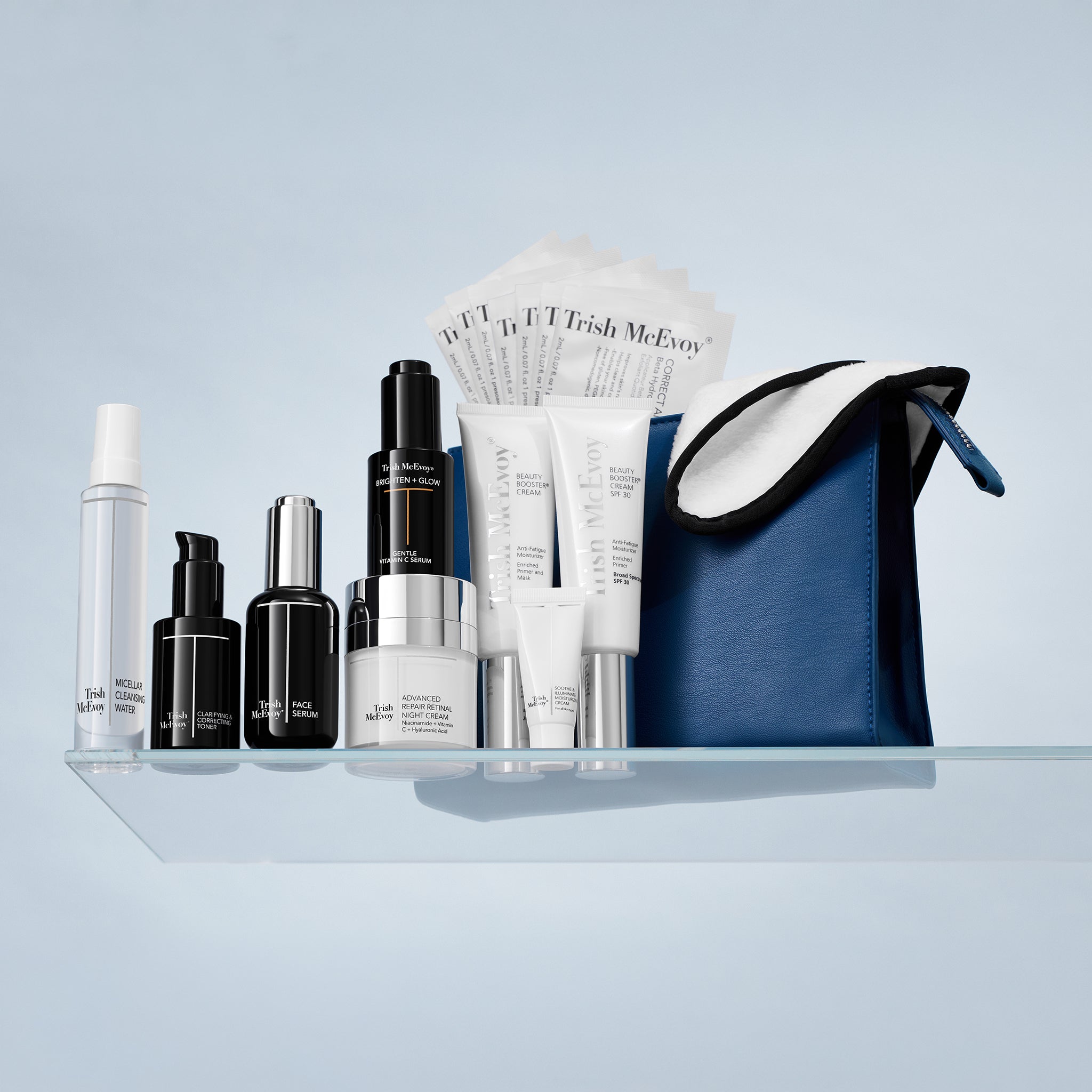A skincare gift set arranged on a clear glass shelf against a pale blue background, including several black and white bottles and tubes, a folded white paper fan, and a blue zip pouch with a white interior.