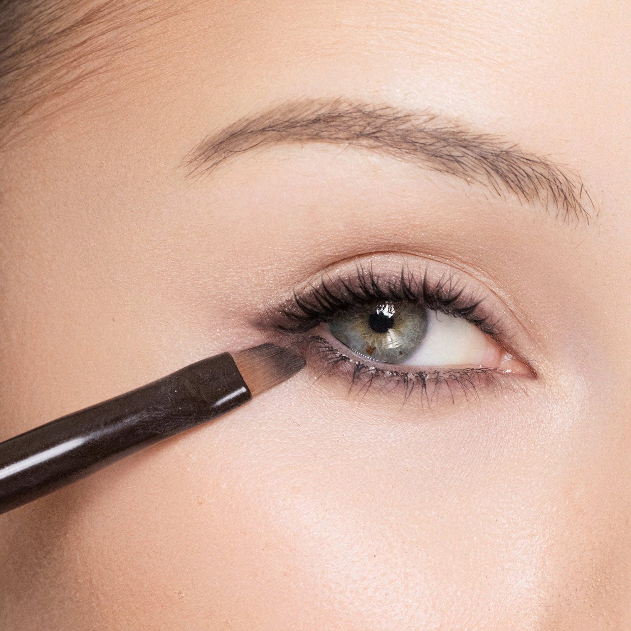 Close-up of a person applying color deep aubergine eye definer with a brush near their eye.