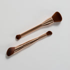 Two makeup brushes with rose gold handles on a light gray background