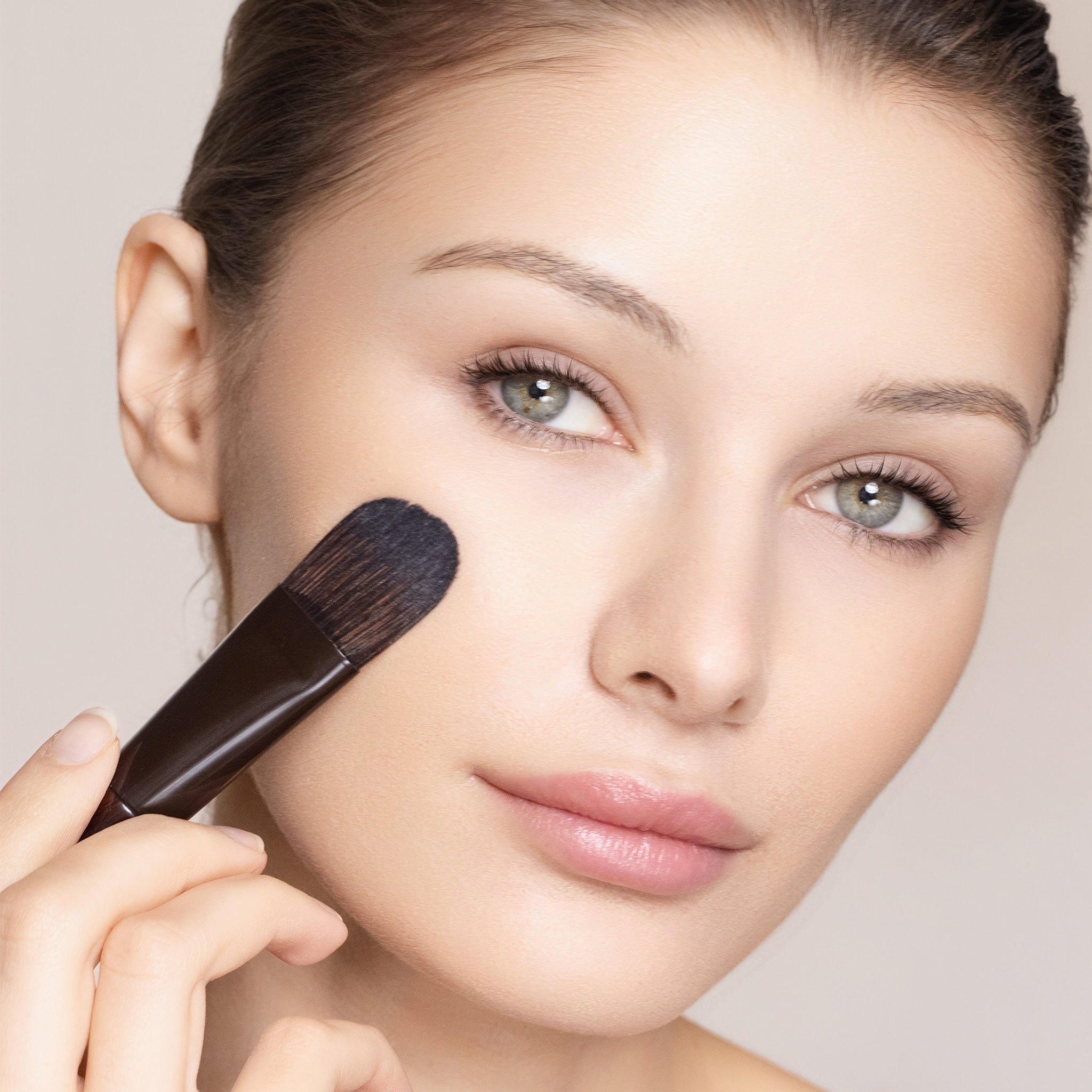 Woman applying makeup with a brush on a neutral background